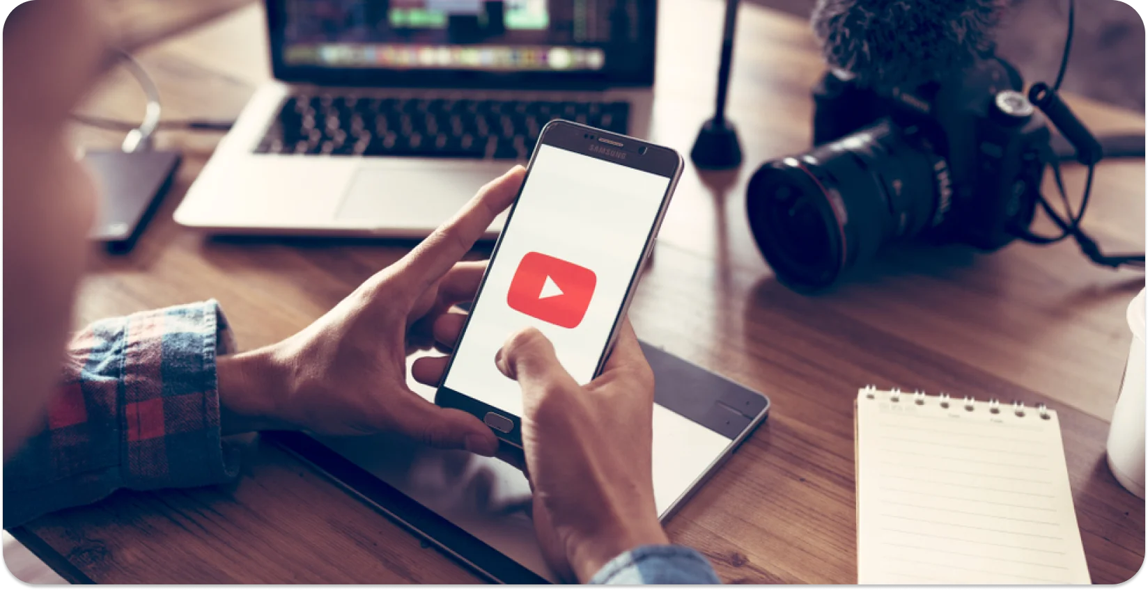 Content creator using a phone with a YouTube play button, with video editing equipment in the background.